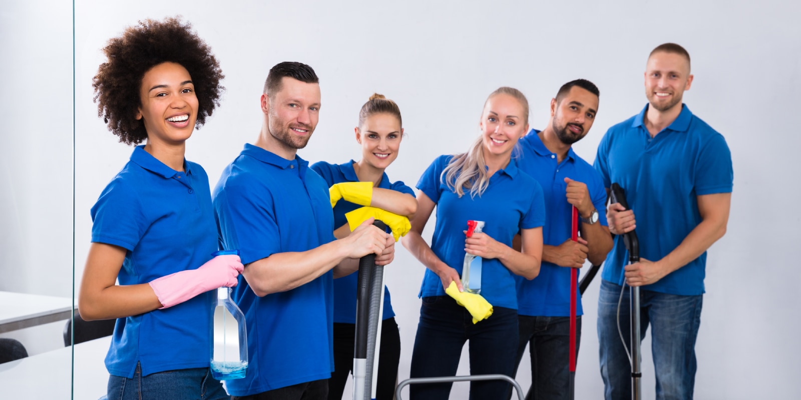 Group of six cleaners in blue shirts smiling and posing with cleaning tools and supplies in a bright room (mops, spray bottles, gloves).